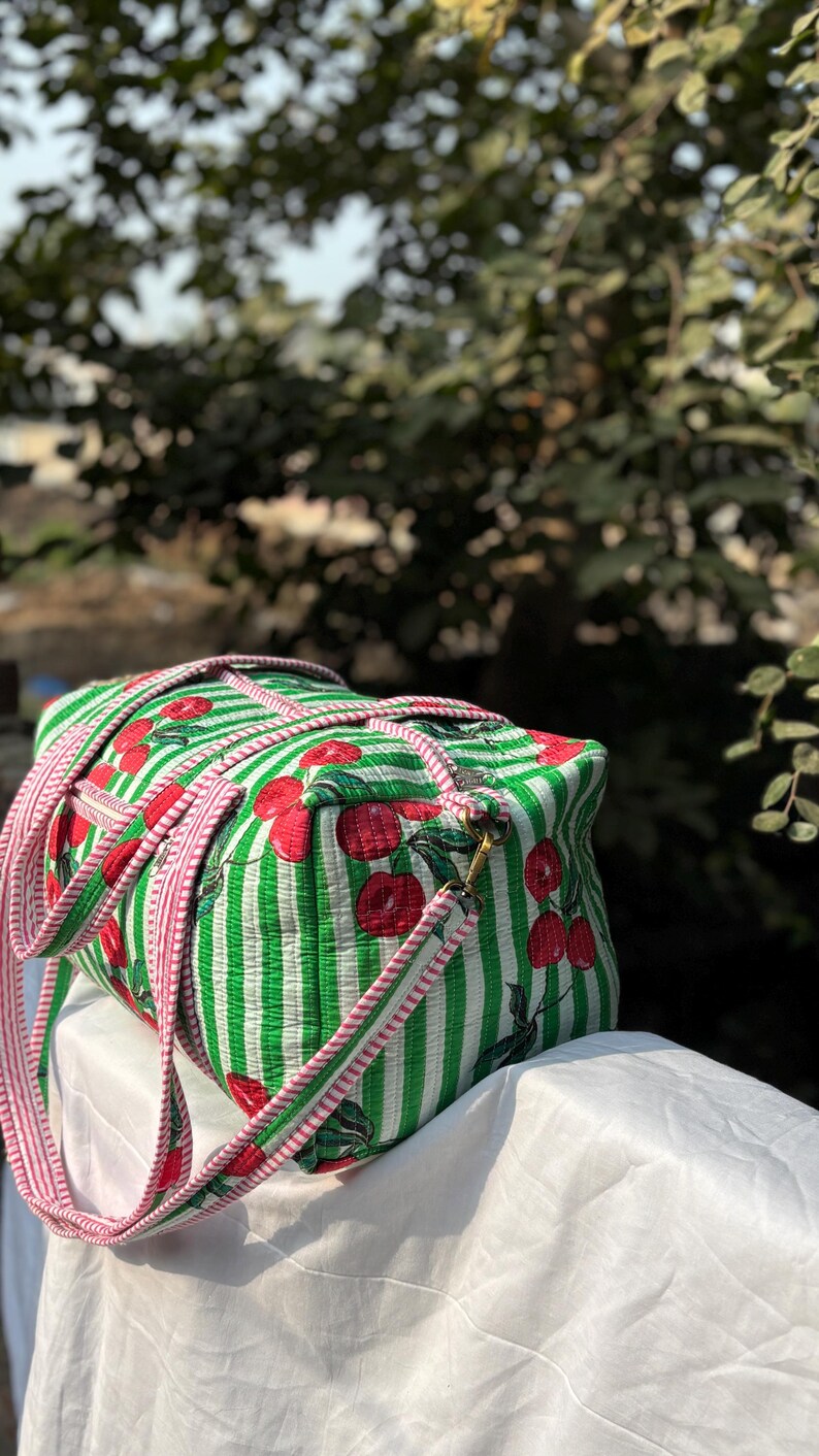 Puede incluir: Una bolsa de lona a rayas verdes y blancas con un estampado de cerezas. La bolsa tiene una correa a rayas rosas y blancas y una cremallera dorada. La bolsa est&aacute; sobre una superficie blanca.
