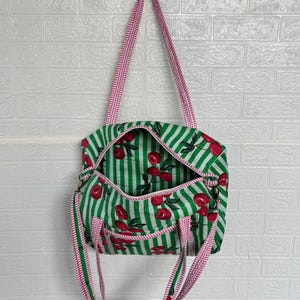 Puede incluir: Bolso tote a rayas verdes y blancas con estampado de cerezas. El bolso tiene una correa a rayas rosas y blancas y un interior a juego. El bolso est&aacute; abierto, revelando el estampado de cerezas en el interior.