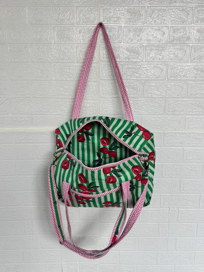 Puede incluir: Bolso tote a rayas verdes y blancas con estampado de cerezas. El bolso tiene una correa a rayas rosas y blancas y un interior a juego. El bolso est&aacute; abierto, revelando el estampado de cerezas en el interior.