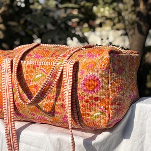 Puede incluir: Bolso tote con estampado floral naranja y rosa con correas a juego. El bolso tiene forma rectangular y está hecho de tela acolchada. Está colocado sobre una superficie blanca.