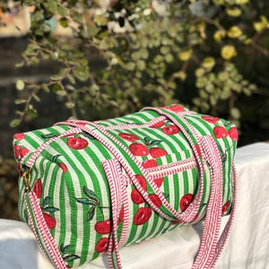 Puede incluir: Una bolsa de lona rectangular a rayas verdes y blancas con un patr&oacute;n de cerezas rojas. La bolsa tiene correas y ribetes a rayas rosas y blancas. El fondo est&aacute; borroso con follaje verde.