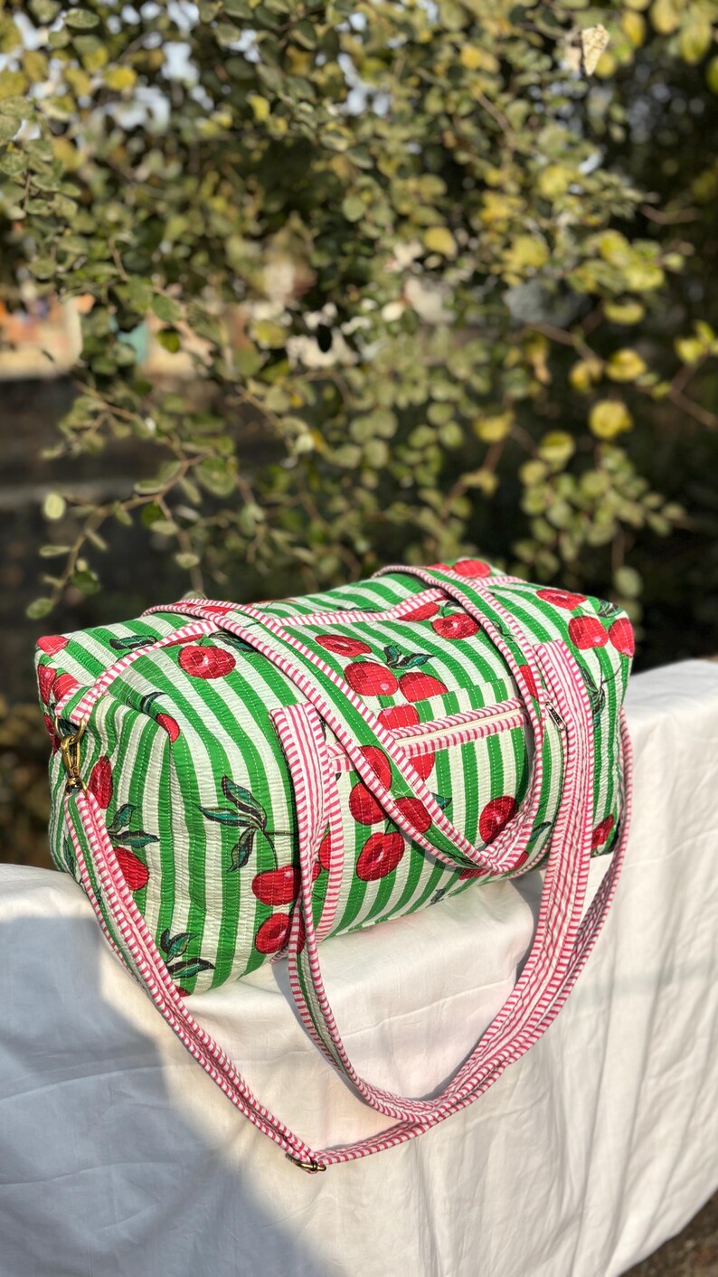 Puede incluir: Una bolsa de lona rectangular a rayas verdes y blancas con un patr&oacute;n de cerezas rojas. La bolsa tiene correas y ribetes a rayas rosas y blancas. El fondo est&aacute; borroso con follaje verde.