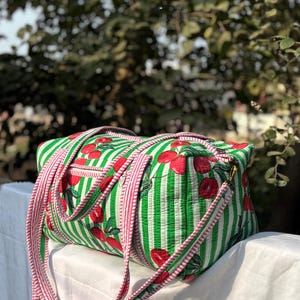 Puede incluir: Una bolsa de lona a rayas verdes y blancas con un estampado de cerezas rojas. La bolsa tiene correas rosas y un cierre de cremallera. La bolsa est&aacute; sobre una superficie blanca.