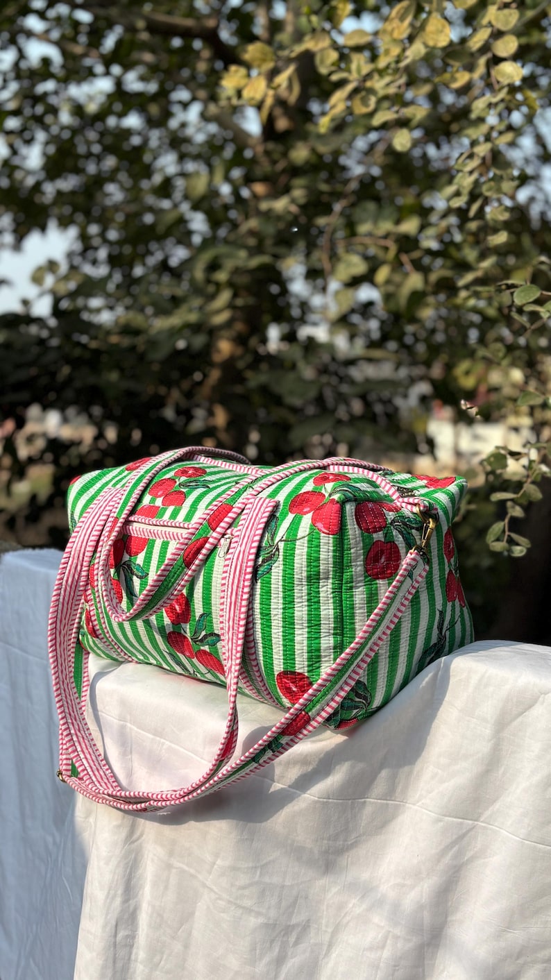 Puede incluir: Una bolsa de lona a rayas verdes y blancas con un estampado de cerezas rojas. La bolsa tiene correas rosas y un cierre de cremallera. La bolsa est&aacute; sobre una superficie blanca.