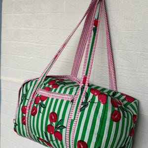Puede incluir: Una bolsa de lona a rayas verdes y blancas con un estampado de cerezas. La bolsa tiene ribetes y correas rosas. Cuenta con un bolsillo con cremallera y est&aacute; colgada contra una pared de ladrillos blancos.