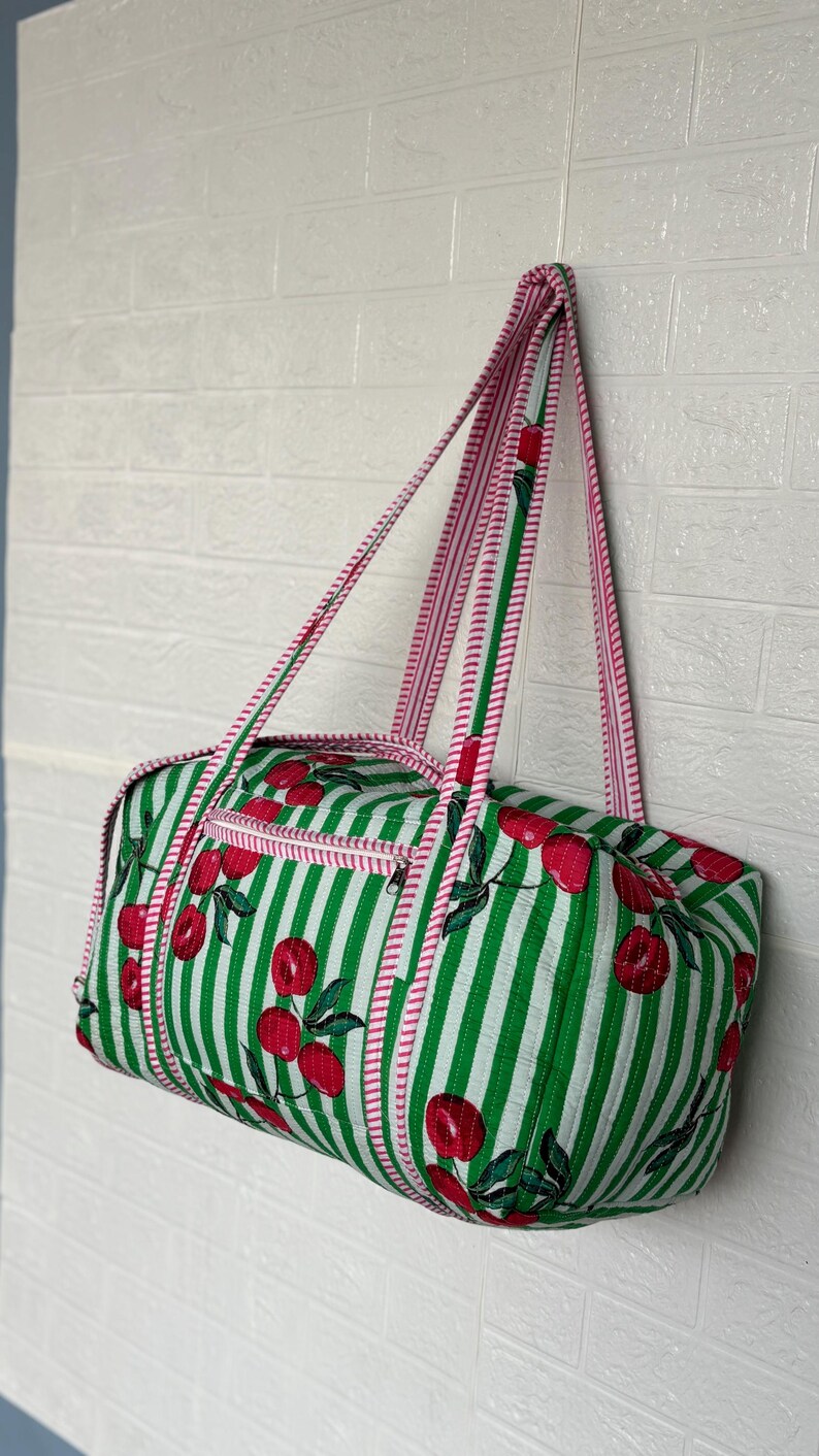Puede incluir: Una bolsa de lona a rayas verdes y blancas con un estampado de cerezas. La bolsa tiene ribetes y correas rosas. Cuenta con un bolsillo con cremallera y est&aacute; colgada contra una pared de ladrillos blancos.