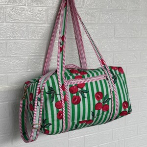 Puede incluir: Una bolsa de lona a rayas verdes y blancas con un estampado de cerezas. La bolsa tiene ribetes y correas rosas. La bolsa cuelga contra una pared de ladrillos blancos.
