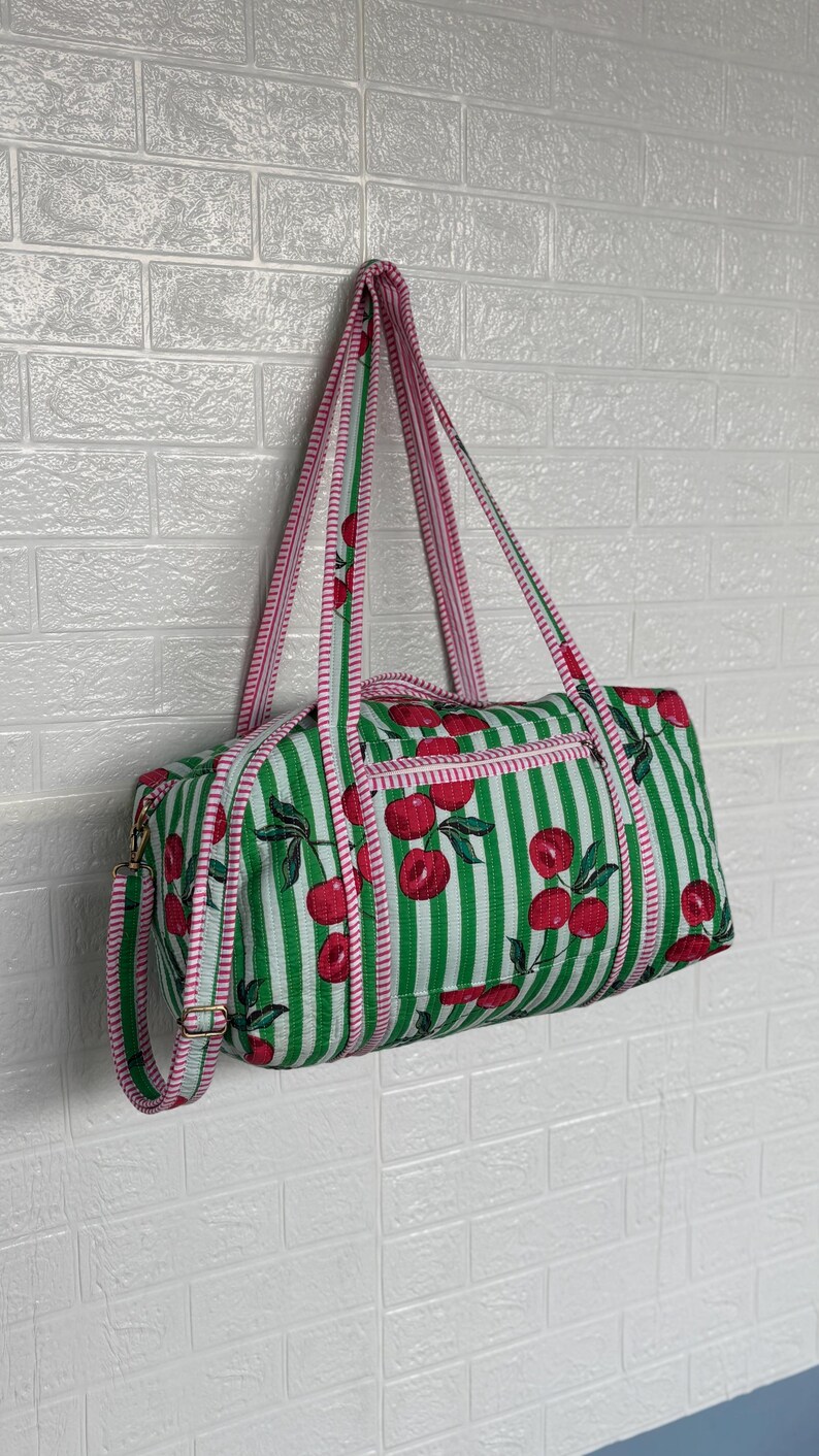 Puede incluir: Una bolsa de lona a rayas verdes y blancas con un estampado de cerezas. La bolsa tiene ribetes y correas rosas. La bolsa cuelga contra una pared de ladrillos blancos.