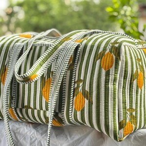 Puede incluir: Una bolsa de viaje acolchada rectangular con un estampado de limones amarillos y hojas verdes sobre un fondo de rayas blancas y verdes. La bolsa incluye dos asas de tela a juego y una forma estructurada con costuras acolchadas visibles.