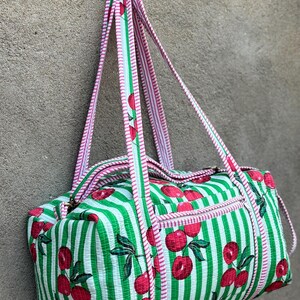 Puede incluir: Bolsa de deporte cil&iacute;ndrica con estampado de rayas verdes y blancas y motivos de cerezas rojas. Cuenta con un bolsillo con cremallera y asas a rayas rosas y blancas. Mide unos 40 cm de largo.