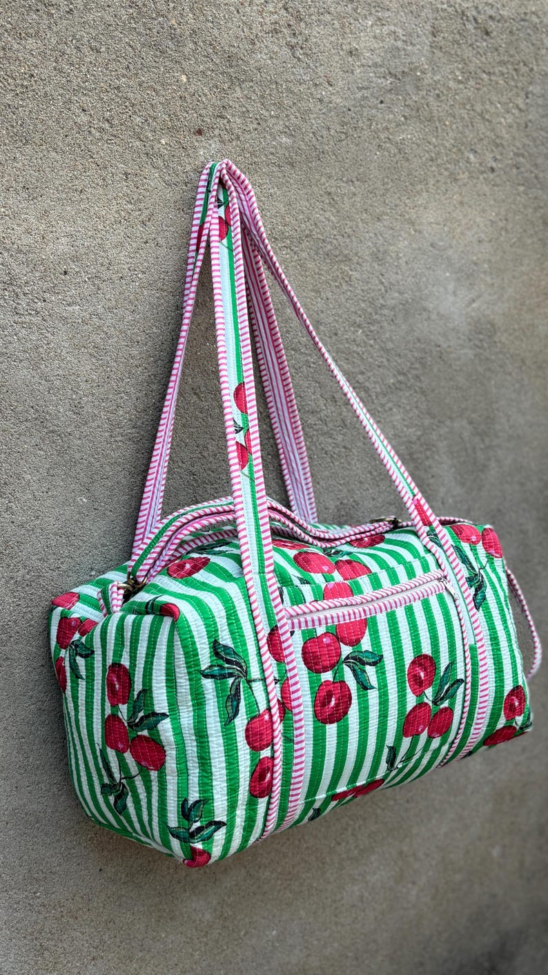 Puede incluir: Bolsa de deporte cil&iacute;ndrica con estampado de rayas verdes y blancas y motivos de cerezas rojas. Cuenta con un bolsillo con cremallera y asas a rayas rosas y blancas. Mide unos 40 cm de largo.