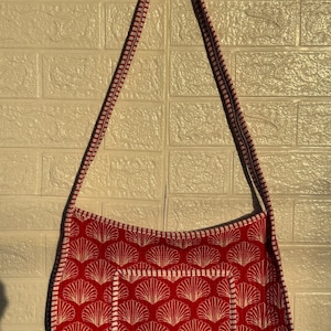 May include: Red shoulder bag with a white seashell pattern. The bag has a matching red and white strap and trim. The bag is hanging against a white brick wall.