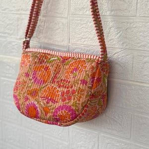 May include: A small, quilted crossbody bag with a floral pattern in shades of orange, pink, and green. The bag has a matching strap and a pink and white striped trim. The bag is hanging against a white brick wall.