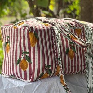 Puede incluir: Una bolsa de viaje acolchada rectangular con un estampado de limones amarillos y hojas verdes sobre un fondo de rayas verticales granates y blancas. La bolsa incluye dos asas de tela a juego y un cierre de cremallera en la parte superior.