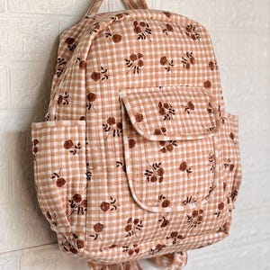 Puede incluir: Una mochila acolchada con un patrón de cuadros vichy en color melocotón claro y blanco, con un pequeño estampado floral marrón. La bolsa incluye un asa superior, dos correas para los hombros, un bolsillo frontal con solapa y dos bolsillos laterales.