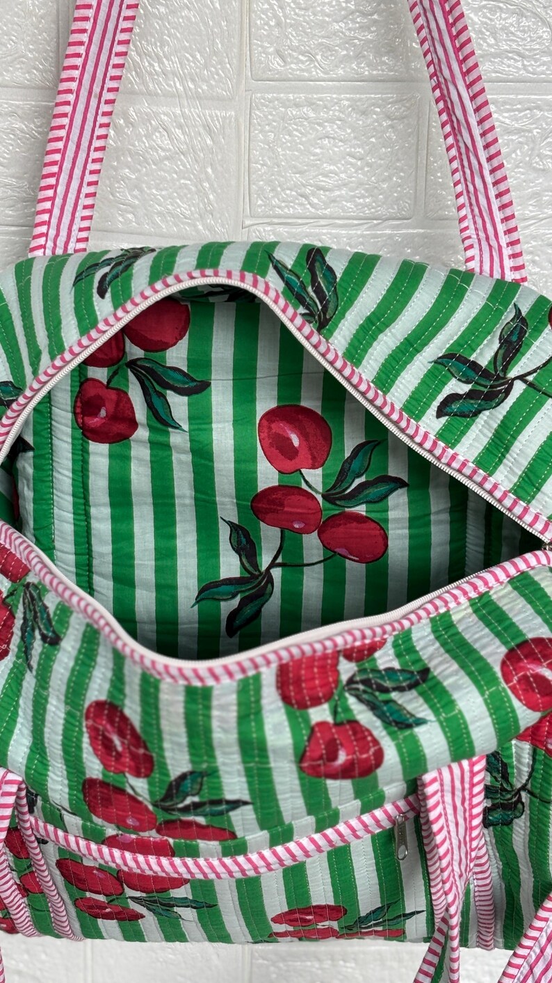 Puede incluir: Bolso tote acolchado con un estampado de rayas verdes y blancas. El bolso presenta un estampado de cerezas en el interior y el exterior. Las correas y los ribetes son de rayas rosas y blancas. El bolso tiene cierre de cremallera.