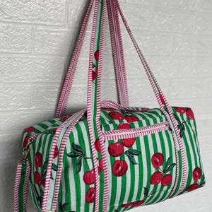 Puede incluir: Bolsa de lona a rayas verdes y blancas con un estampado de cerezas. La bolsa tiene ribetes y correas rosas y una cremallera dorada. La bolsa est&aacute; colgada contra una pared de ladrillos blancos.