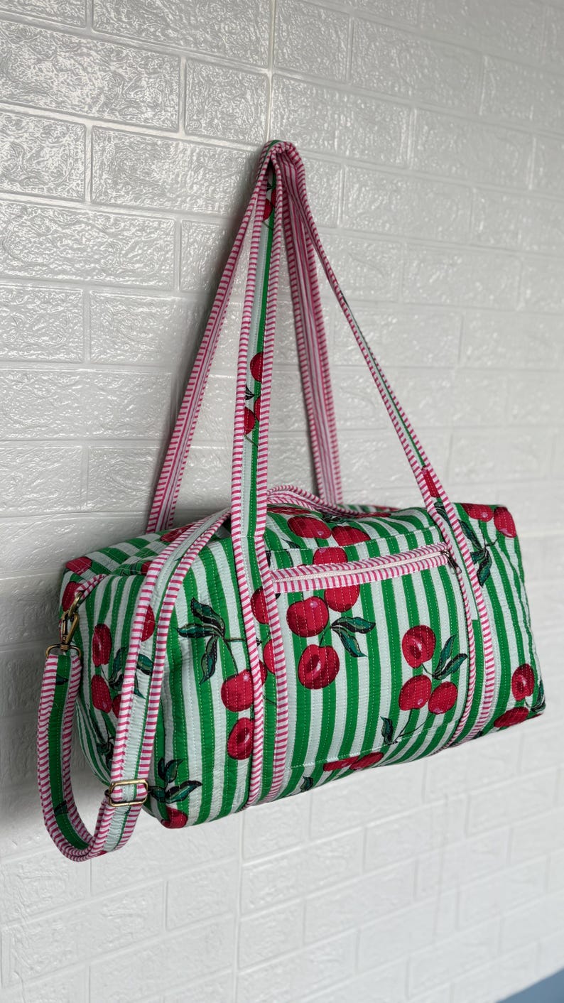Puede incluir: Bolsa de lona a rayas verdes y blancas con un estampado de cerezas. La bolsa tiene ribetes y correas rosas y una cremallera dorada. La bolsa est&aacute; colgada contra una pared de ladrillos blancos.