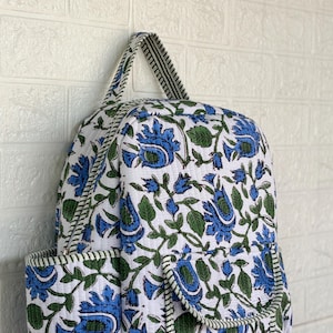 May include: A quilted backpack featuring a white base with a repeating floral pattern in blue and green. The bag includes a top handle, two side pockets, and a front pocket with a flap, all accented with a green and white striped fabric trim.