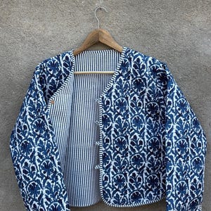 May include: A blue and white patterned jacket with a floral design. The jacket is open, revealing a striped lining. It hangs on a wooden hanger against a neutral background.