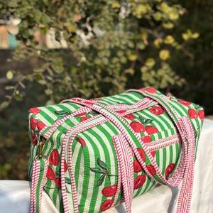 Puede incluir: Una bolsa de lona rectangular a rayas verdes y blancas con patrones de cerezas rojas. La bolsa tiene correas a rayas rojas y blancas y herrajes met&aacute;licos dorados. El fondo est&aacute; borroso con follaje verde.
