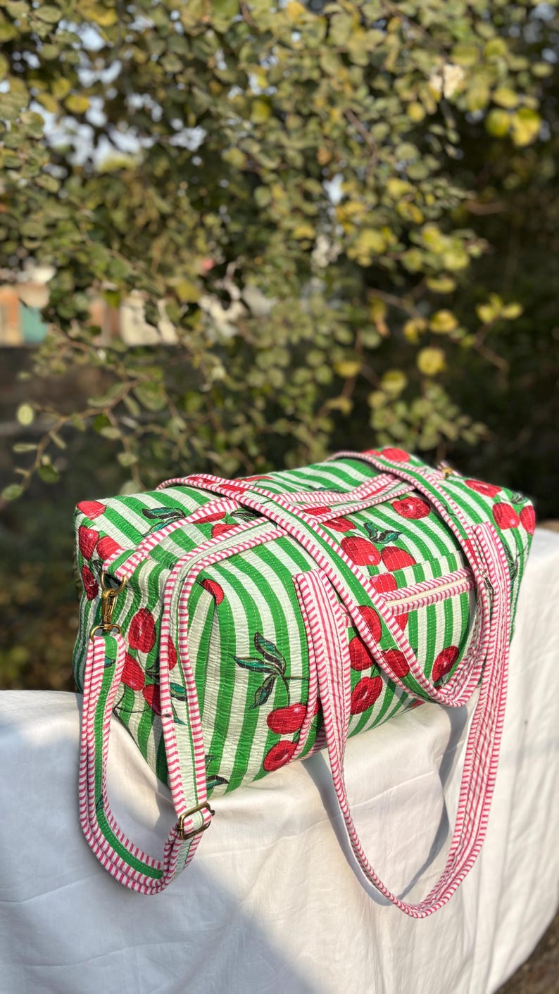 Puede incluir: Una bolsa de lona rectangular a rayas verdes y blancas con patrones de cerezas rojas. La bolsa tiene correas a rayas rojas y blancas y herrajes met&aacute;licos dorados. El fondo est&aacute; borroso con follaje verde.