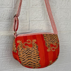 Op de afbeelding: Rode en oranje crossbodytas met een tijgerprint. De tas heeft een gestreepte band en een ritssluiting. De tas is gemaakt van een gewatteerde stof.