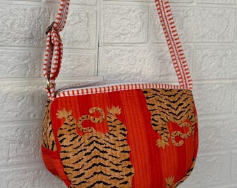 May include: A red and orange crossbody bag with a tiger print design. The bag has a striped strap and a zipper closure. The bag is made of a quilted fabric.