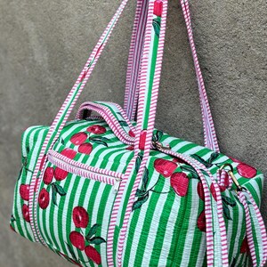 Puede incluir: Una bolsa de lona a rayas verdes y blancas con un estampado de cerezas. La bolsa tiene ribetes y correas rosas. La bolsa tiene un bolsillo con cremallera y herrajes dorados. La bolsa est&aacute; hecha de tela acolchada.