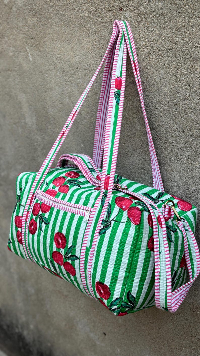 Puede incluir: Una bolsa de lona a rayas verdes y blancas con un estampado de cerezas. La bolsa tiene ribetes y correas rosas. La bolsa tiene un bolsillo con cremallera y herrajes dorados. La bolsa est&aacute; hecha de tela acolchada.