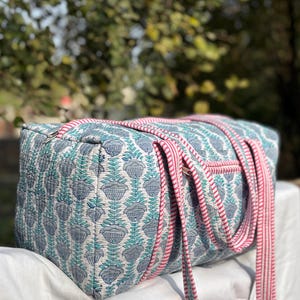 Puede incluir: Bolso de viaje acolchado con un estampado floral en tonos azules y turquesas sobre un fondo blanco. El bolso tiene asas y ribetes a rayas rosas y blancas. El bolso es rectangular y parece ser de algodón.