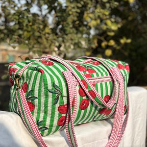 Puede incluir: Una bolsa de lona a rayas verdes y blancas con un estampado de cerezas. La bolsa tiene una correa y ribete rosas, y un peque&ntilde;o bolsillo lateral. La bolsa est&aacute; sobre una superficie blanca.