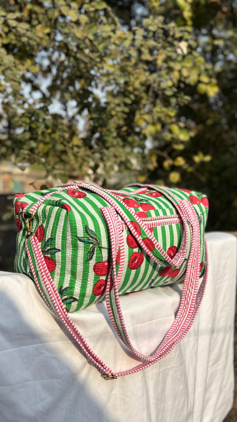 Puede incluir: Una bolsa de lona a rayas verdes y blancas con un estampado de cerezas. La bolsa tiene una correa y ribete rosas, y un peque&ntilde;o bolsillo lateral. La bolsa est&aacute; sobre una superficie blanca.