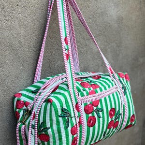 Puede incluir: Una bolsa de lona a rayas verdes y blancas con un estampado de cerezas. La bolsa tiene ribetes y correas rosas. La bolsa cuelga contra una pared gris.