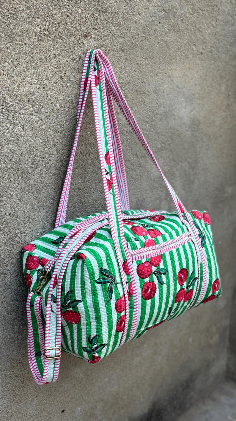 Puede incluir: Una bolsa de lona a rayas verdes y blancas con un estampado de cerezas. La bolsa tiene ribetes y correas rosas. La bolsa cuelga contra una pared gris.