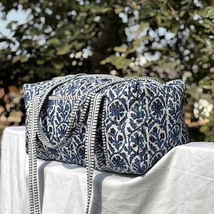 Puede incluir: Un bolso tote rectangular con un estampado floral azul y blanco. El bolso tiene correas largas y está sobre una superficie blanca. El fondo está borroso con follaje verde.