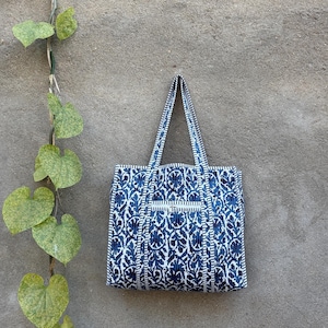 May include: A blue and white patterned tote bag with matching handles. The bag features a floral design and a small pocket. The bag is hanging against a gray wall with green leaves.