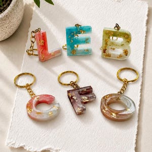 May include: Six resin letter keychains featuring gold leaf flakes embedded within the material. The letters L, E, B, C, F, and O are displayed, each attached to a gold-toned metal split ring and a short chain link for secure attachment.