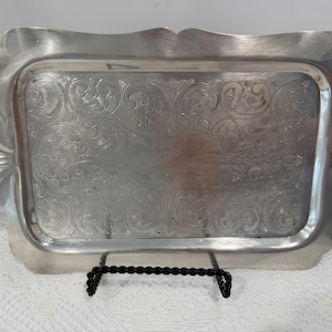 May include: A silver-coloured rectangular tray with embossed floral patterns. The tray has a scalloped edge design and is displayed on a black metal stand. The tray is likely used for serving or display purposes.