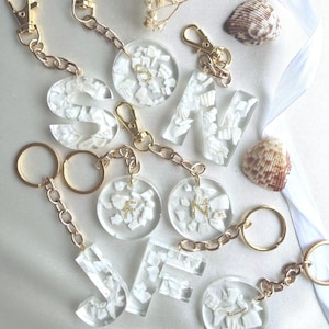 May include: A collection of clear resin keychains featuring embedded white shell fragments and gold-toned metal hardware. The keychains include various letter shapes and circular discs, some with gold glitter letters S, F, and N visible inside.