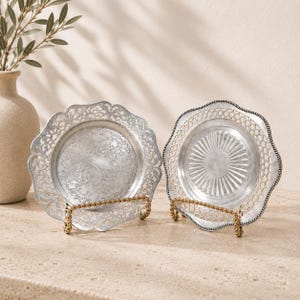 May include: Two silver-toned metal decorative plates displayed on gold-toned wire stands. The left plate features an intricate filigree border and a textured centre, while the right plate has a scalloped edge with a lattice pattern and a ribbed centre.