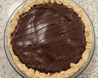 Puede incluir: Una tarta de chocolate redonda en un plato de cristal transparente que presenta un centro de ganache de chocolate negro suave con un patrón de remolinos decorativo y un borde grueso de glaseado color canela alrededor del postre.
