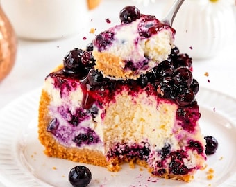 Puede incluir: Una porción de tarta de queso con arándanos en un plato blanco con una cuchara de plata levantando un trozo. El postre presenta una base de galleta dorada, un relleno blanco cremoso con bayas oscuras y una cobertura de arándanos.