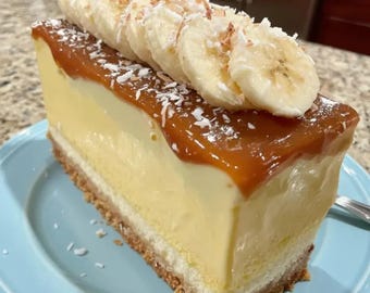 Puede incluir: Una porción rectangular de tarta de queso con plátano sobre un plato azul claro. El postre presenta una base dorada, una capa gruesa de queso crema amarillo pálido, una cobertura de caramelo suave y rodajas de plátano espolvoreadas con coco.