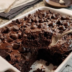 Op de afbeelding: Een witte rechthoekige bakvorm gevuld met chocoladetaart, bedekt met chocolade glazuur en chocolade chips. Een stuk cake wordt opgetild met een zilveren vork. Een vork en servet zijn links, en een bord en glas zijn op de achtergrond.