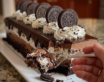 Puede incluir: Un pastel rectangular de chocolate con capas de crema blanca y ganache de chocolate oscuro, cubierto con remolinos de crema batida y galletas sándwich de chocolate enteras. Una cuchara de plata levanta una porción del pastel.