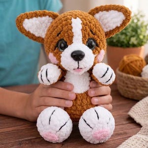 May include: A plush crochet toy shaped like a corgi dog with brown and white fur, large black eyes, a black nose, and pink cheeks. The stuffed animal features oversized ears, white paws with black stitched details, and pink accents on the feet.