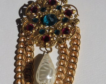 May include: A gold-tone brooch with a floral design and red, green, and blue rhinestones. A single white pearl dangles from the center of the brooch, and a gold chain hangs from the bottom.
