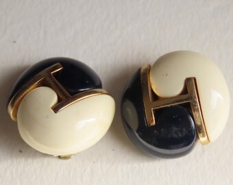 Könnte beinhalten: Ein Paar Vintage-Ohrclips. Jeder Ohrring hat ein zweifarbiges Design mit einem marineblauen und cremefarbenen Halbkreis, akzentuiert mit einer goldfarbenen "H"-Form aus Metall. Die Ohrringe haben ein glänzendes Finish.