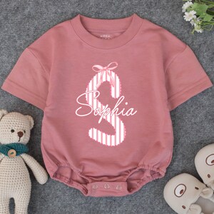 May include: Dusty rose baby romper with the name "Sophia" in a white script font. The letter "S" is decorated with white and pink stripes and a pink bow. The short-sleeved onesie has a round neckline and snap closures.